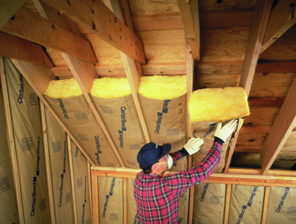 Technician in red plaid shirt, wearing face mask and gloves, installing yellow fiberglass batt insulation in an attic ceiling.